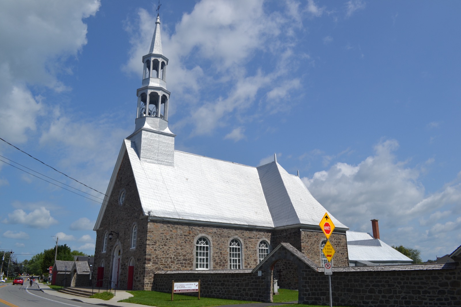 L'église Saint-Mathias en pierre avec son clocher argenté et le mur de son cimetière, en bordure du chemin des Patriotes